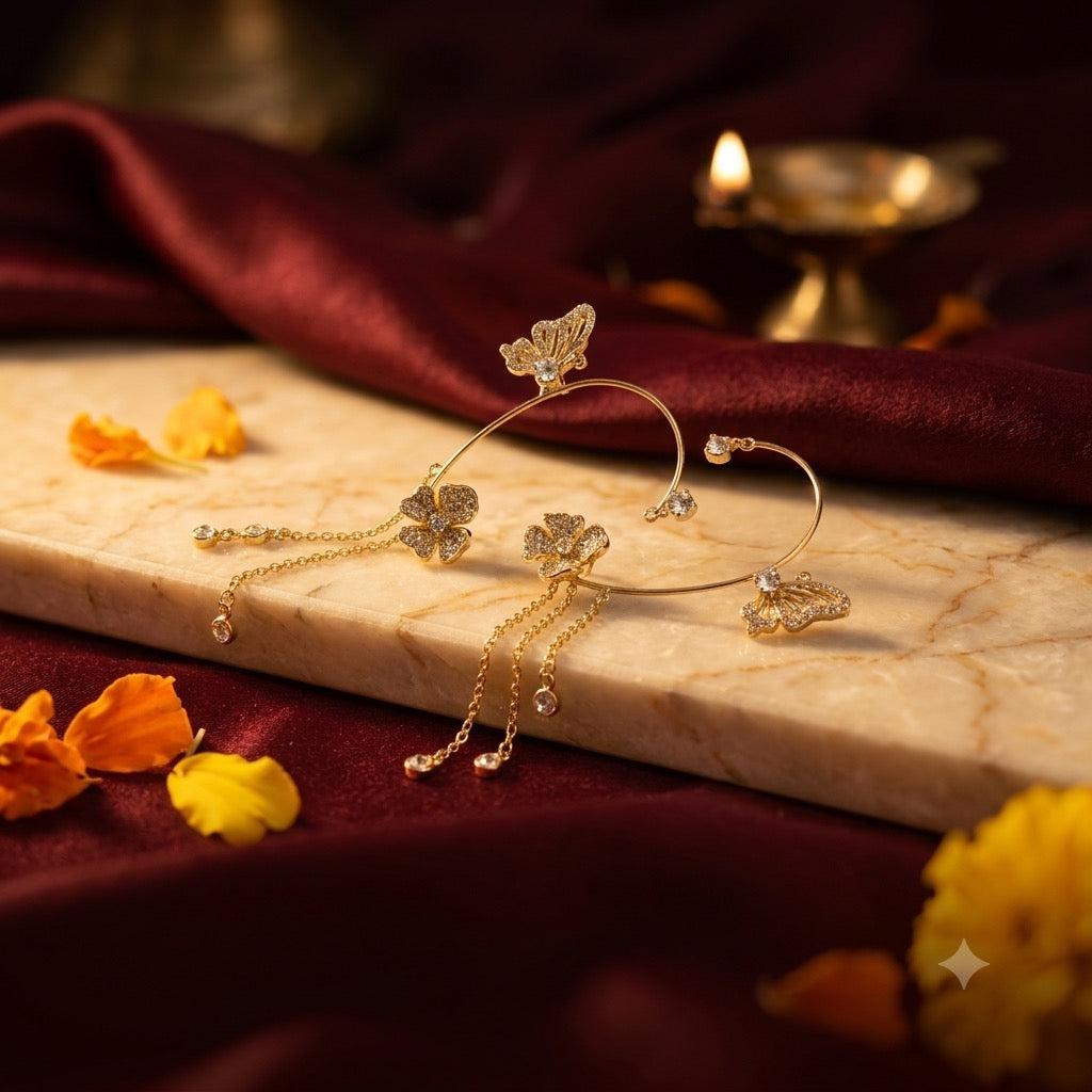 Antique-gold butterfly and floral ear cuffs with delicate chains on cream marble, with deep maroon silk, marigold petals and a lit brass diya
